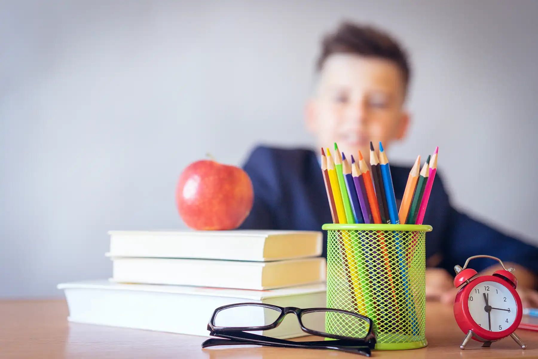 Educational workspace with books, red apple, eyeglasses, colorful pencils in a green holder, and red alarm clock on a desk; blurred person in background