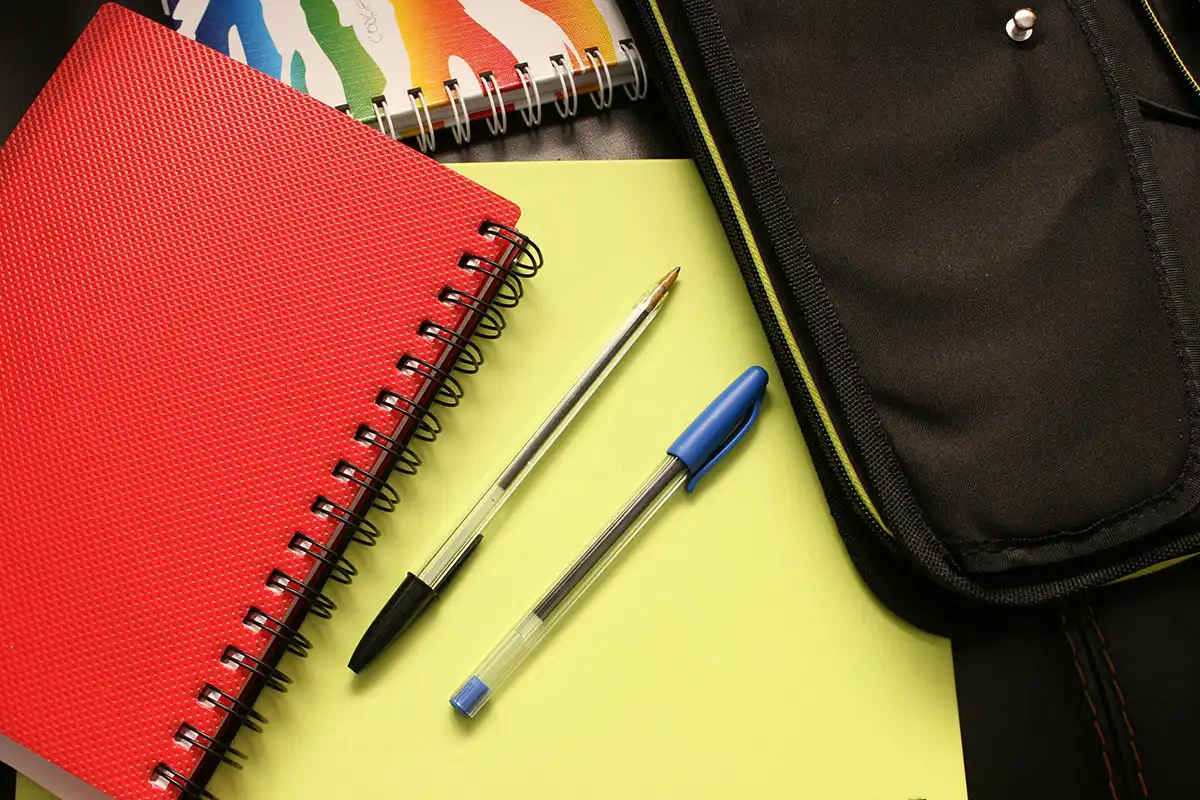 Flat lay of school supplies including a red spiral notebook, black and blue pens, a yellow folder, a multicoloured notepad, and a black pencil case on a desk—symbolising education, learning, and back-to-school readiness.