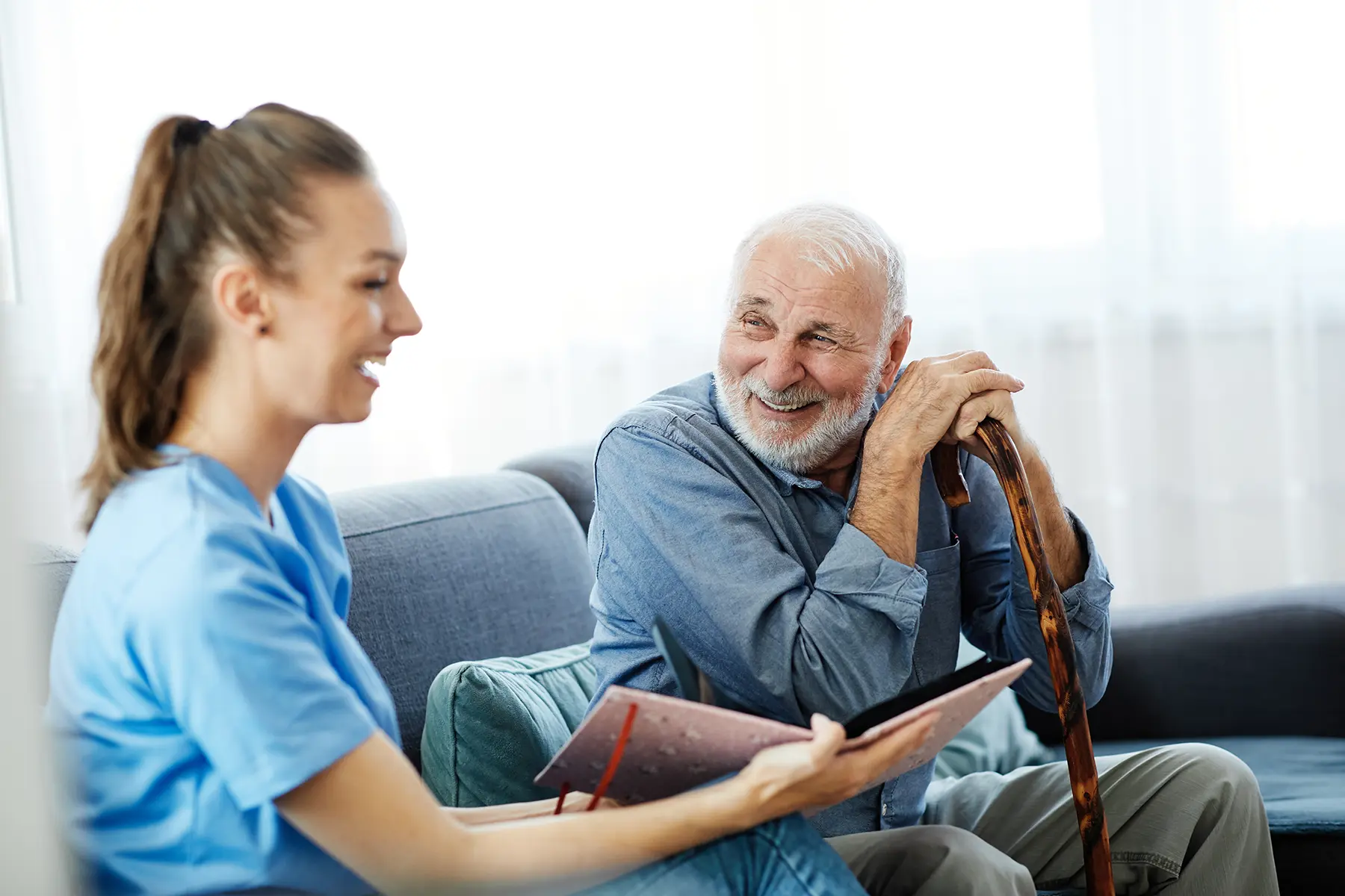 Smiling female care worker sharing a photo album with a happy elderly man holding a wooden cane
