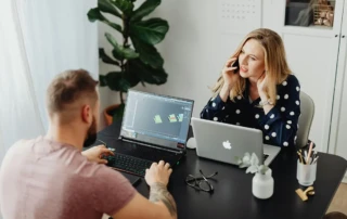 Two colleagues working at a black desk, one man using a laptop with 3D design software open and a woman talking on a mobile phone while working on her laptop. Glasses, stationery, and small plants are on the desk, with a large potted plant in the background.