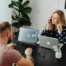 Two colleagues working at a black desk, one man using a laptop with 3D design software open and a woman talking on a mobile phone while working on her laptop. Glasses, stationery, and small plants are on the desk, with a large potted plant in the background.