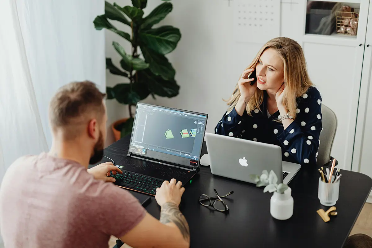 Two colleagues working at a black desk, one man using a laptop with 3D design software open and a woman talking on a mobile phone while working on her laptop. Glasses, stationery, and small plants are on the desk, with a large potted plant in the background.