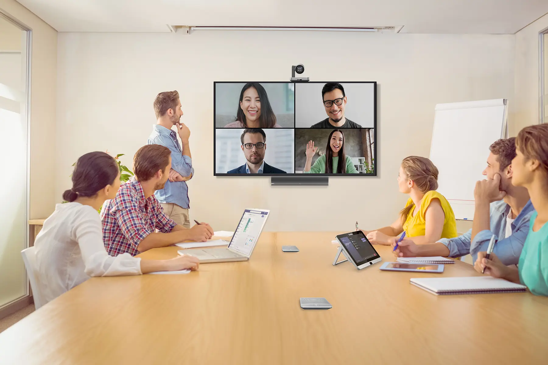 Colleagues sitting around a conference table with laptops and devices, looking at a large screen on the wall with a video conference split screen with 4 participants