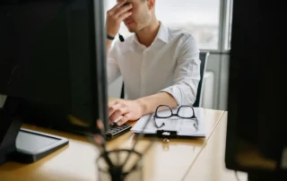 Office worker in a white shirt sitting at a desk with two computer monitors, holding his head in frustration while typing. A clipboard with papers and a pair of glasses rests on the desk in front of him.