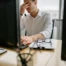 Office worker in a white shirt sitting at a desk with two computer monitors, holding his head in frustration while typing. A clipboard with papers and a pair of glasses rests on the desk in front of him.