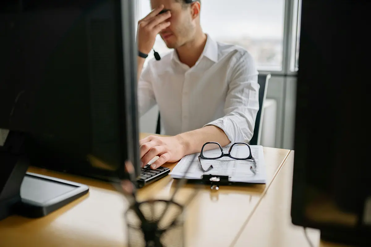 Office worker in a white shirt sitting at a desk with two computer monitors, holding his head in frustration while typing. A clipboard with papers and a pair of glasses rests on the desk in front of him.