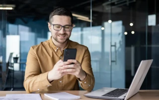 Smiling professional in brown shirt using smartphone at modern office desk with laptop and documents