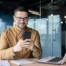 Smiling professional in brown shirt using smartphone at modern office desk with laptop and documents