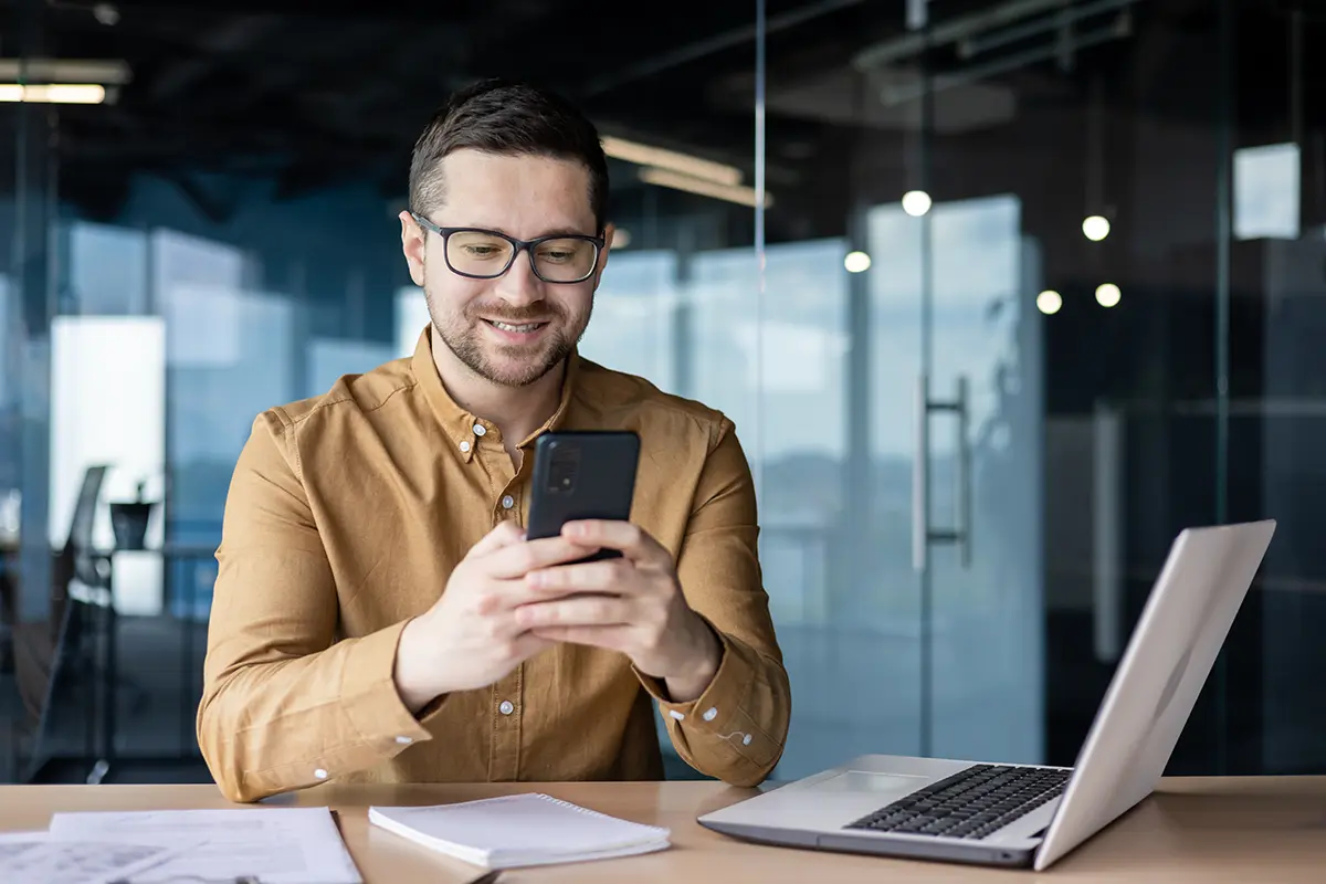 Smiling professional in brown shirt using smartphone at modern office desk with laptop and documents