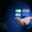 Person in blue shirt holding a smartphone and presenting a digital overlay with toggle switches for years 2025 and 2026, symbolizing future planning and technological transition.