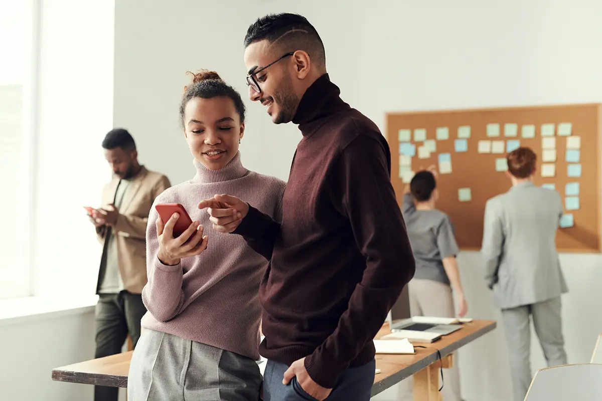 NFP_Cloud-Comms Group of people in an office: two in the foreground interacting with a red smartphone, and two in the background near a corkboard covered in sticky notes — highlighting teamwork, mobile technology, and collaborative planning.
